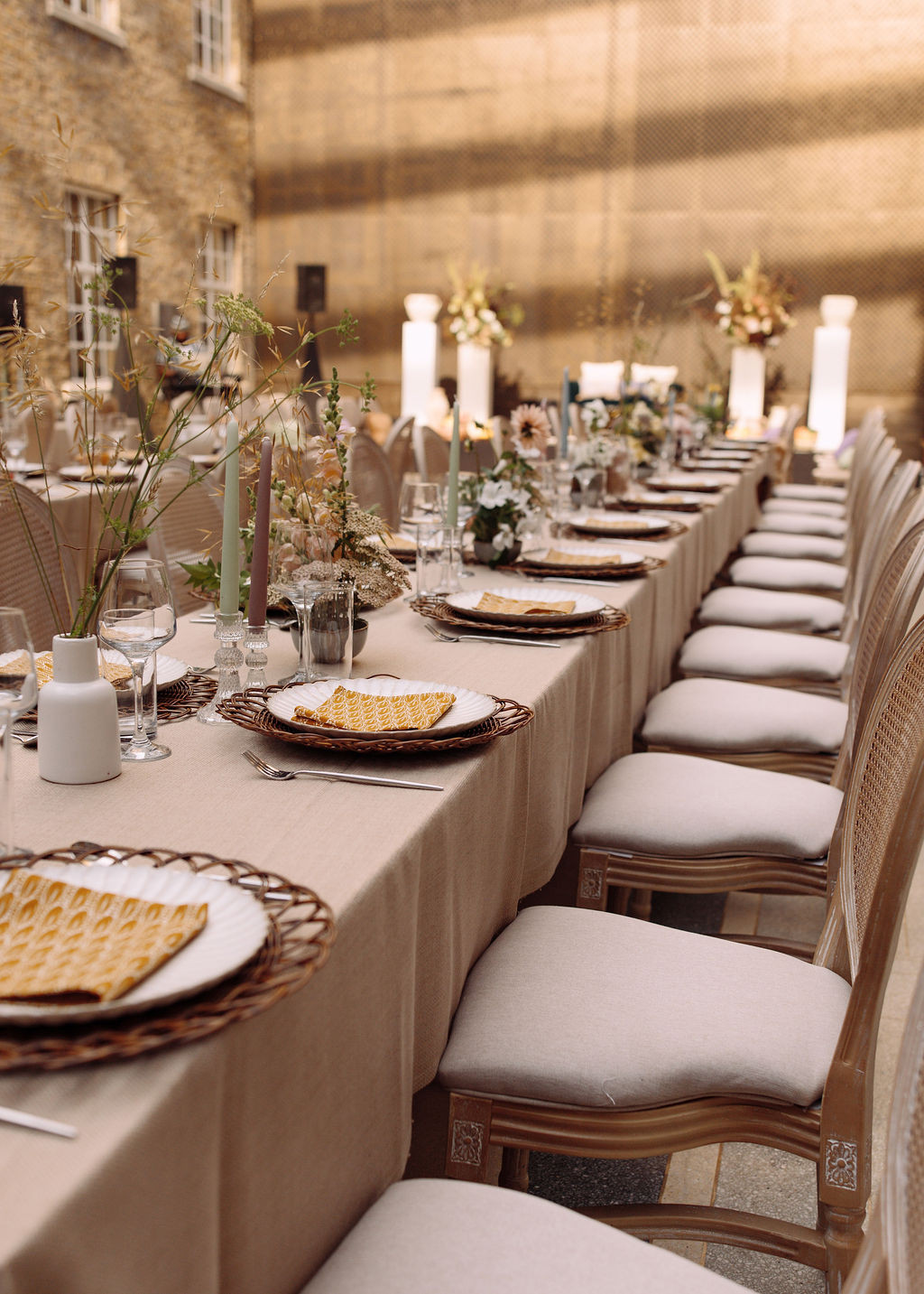 Long linen table with woven chargers and tapered candles against a brick facade in evening light