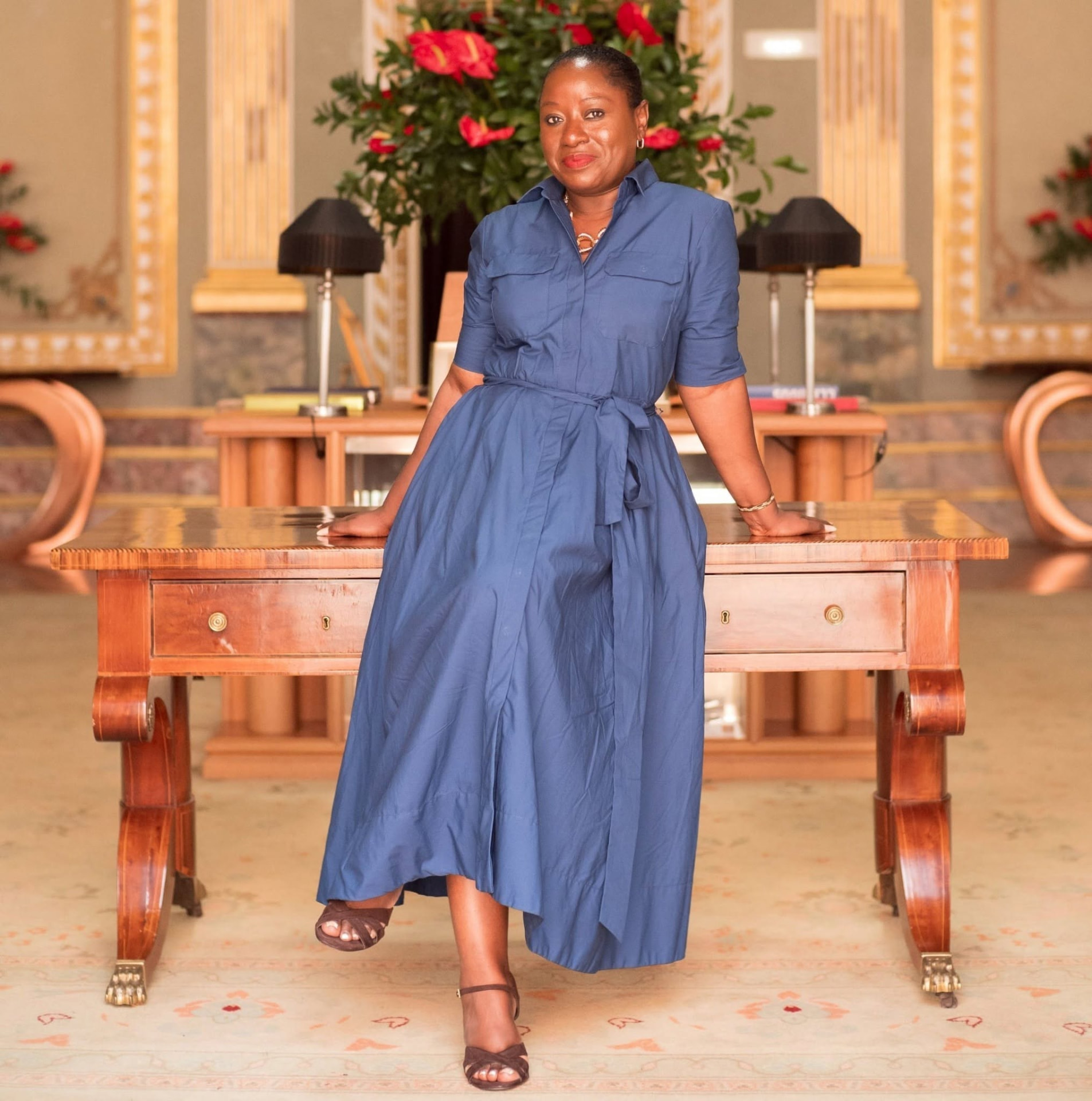 Founder of MWP Collective in a navy shirt dress, seated against an antique writing desk in a gilded state room