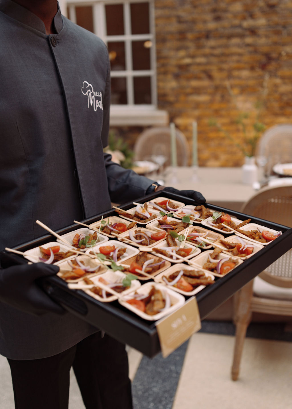 Server in a chef's jacket carrying a tray of suya skewers with red onion and tomato in palm-leaf dishes
