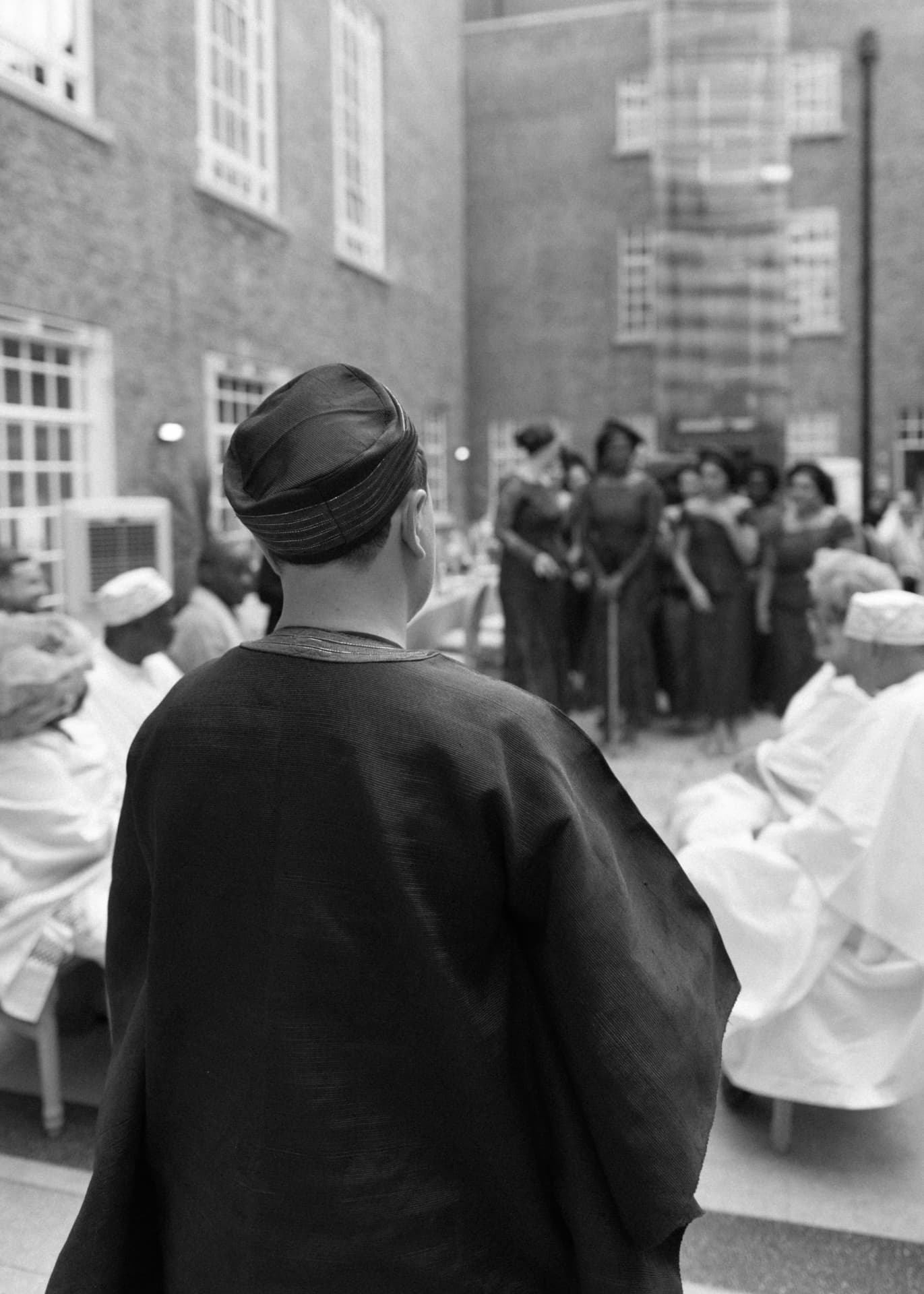 Guest in an agbada and cap watching the ceremony in a brick atrium, black and white