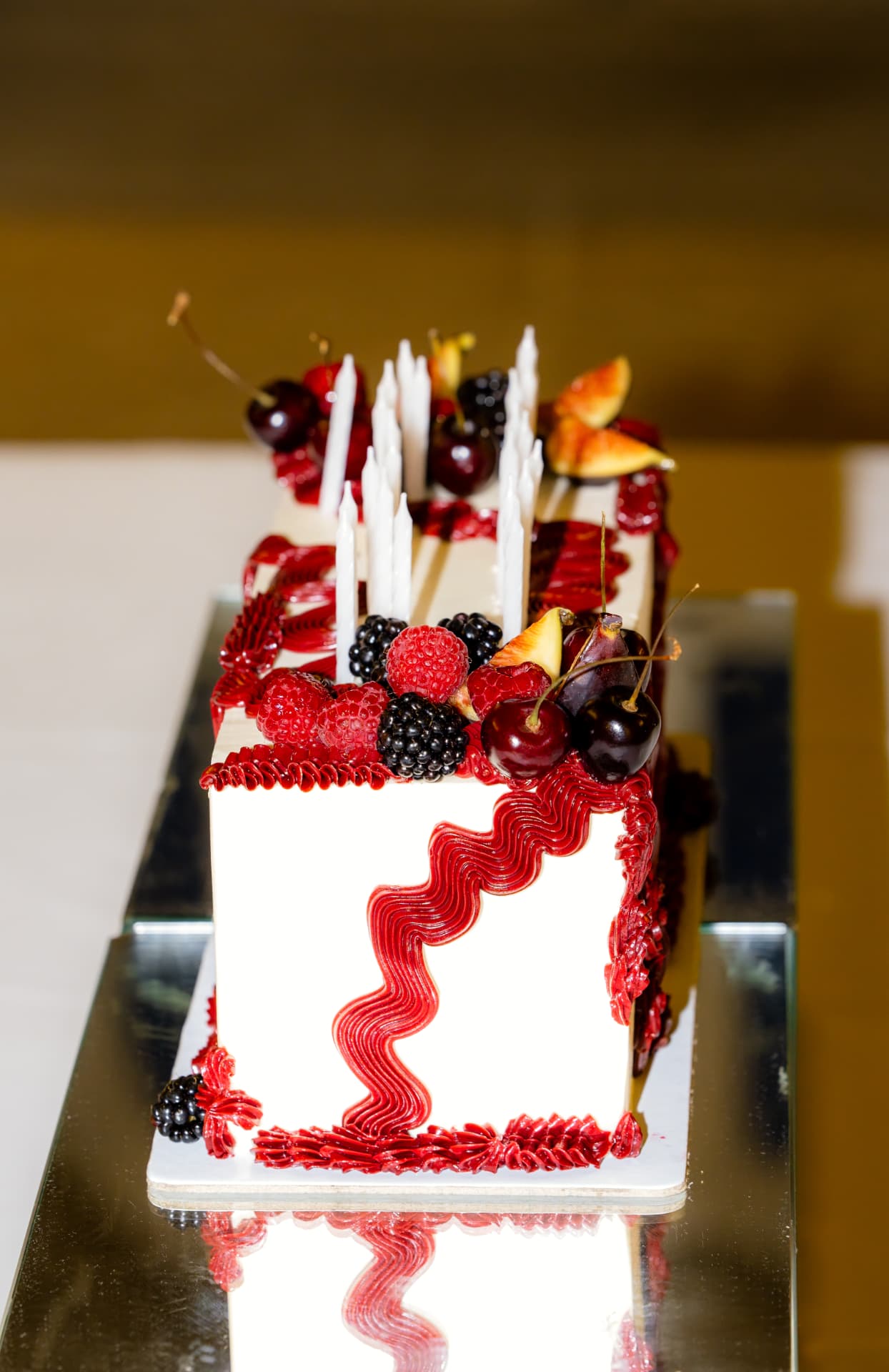 A white celebration cake with red piped scallops, fresh berries, figs and tall candles on a mirrored plinth