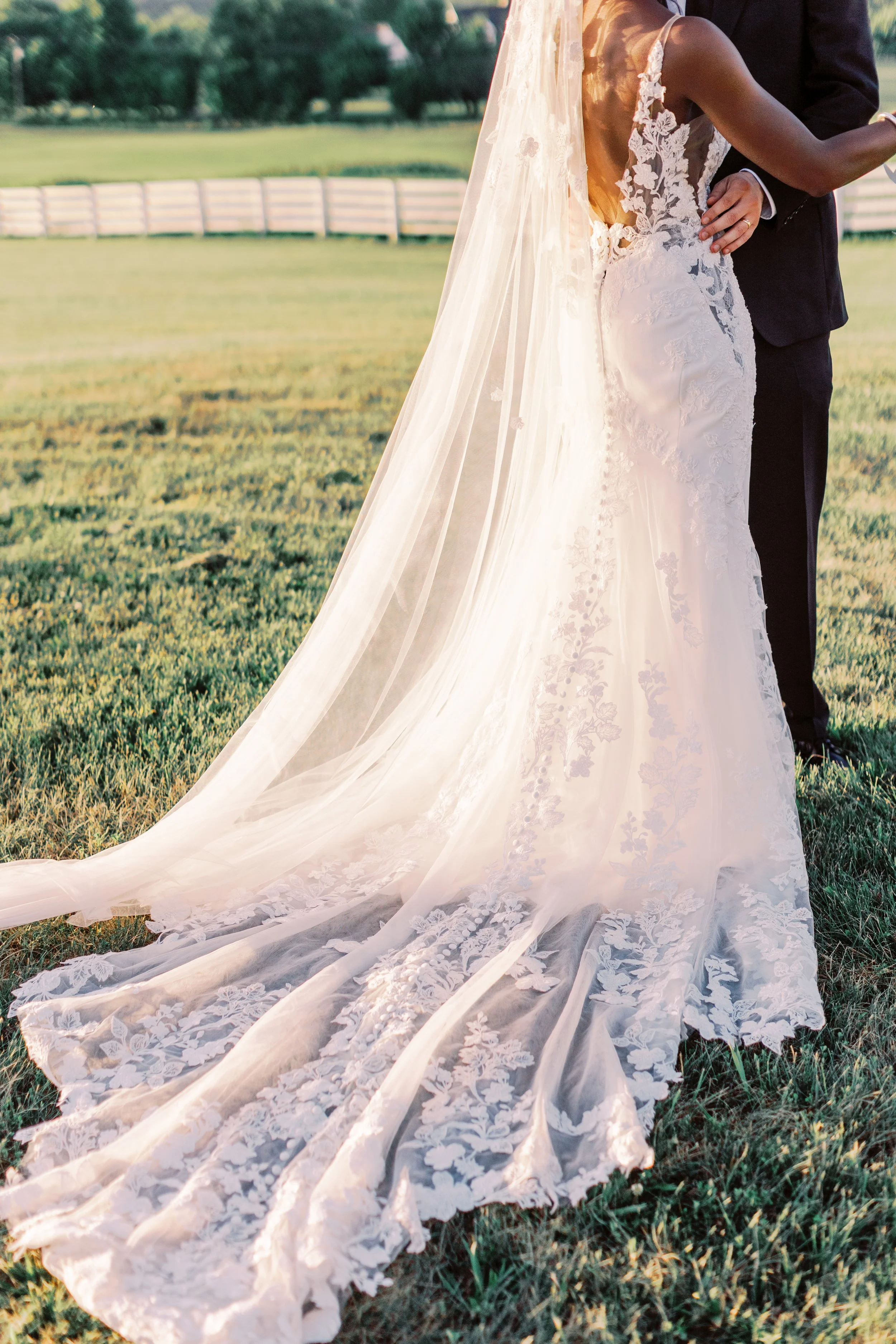 Detail of a corded lace gown and cathedral veil trailing across estate lawn