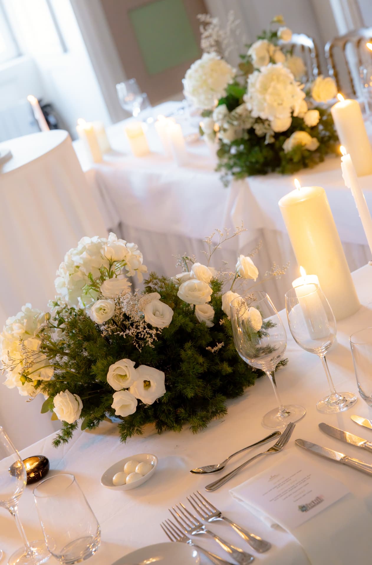 White hydrangeas, roses and lisianthus in a low green arrangement beside pillar candles on a long table