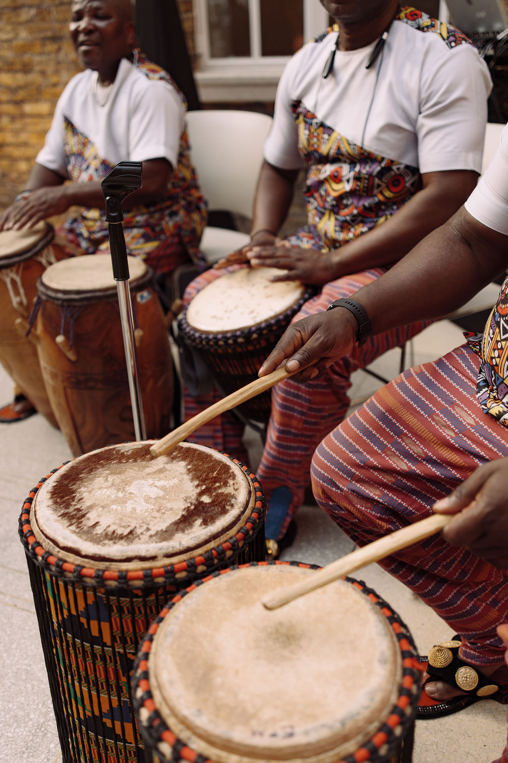 Drummers in white and Ankara cloth playing talking drums and djembes in a brick courtyard