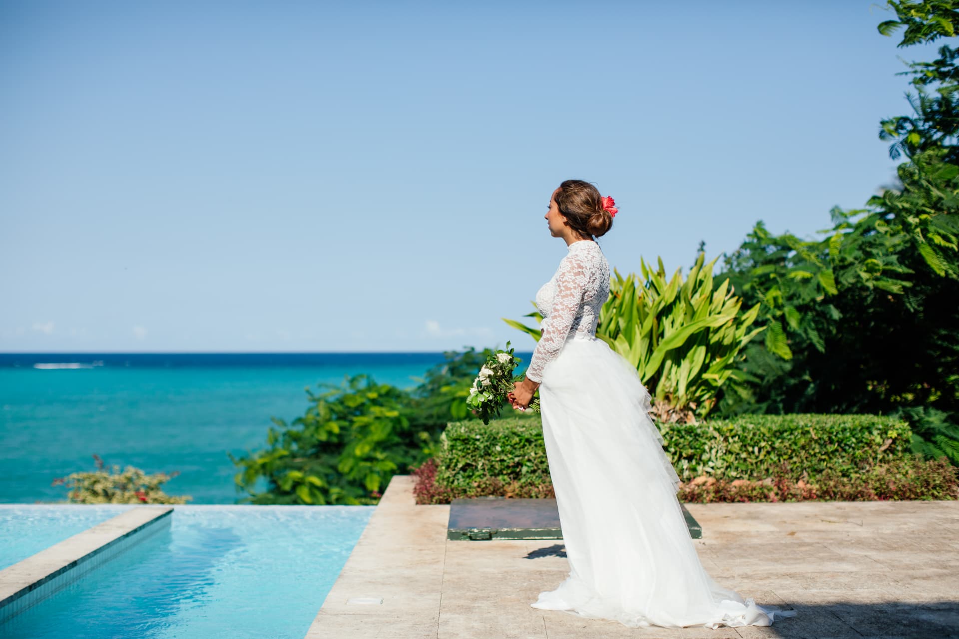 Bride in a lace gown holding a bouquet beside an infinity pool above the Zanzibar coast