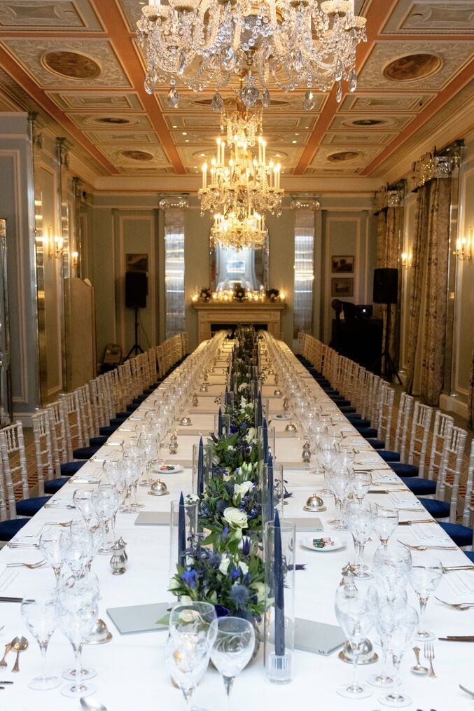 A long banquet table beneath crystal chandeliers in a gilded ballroom, laid with blue and white florals
