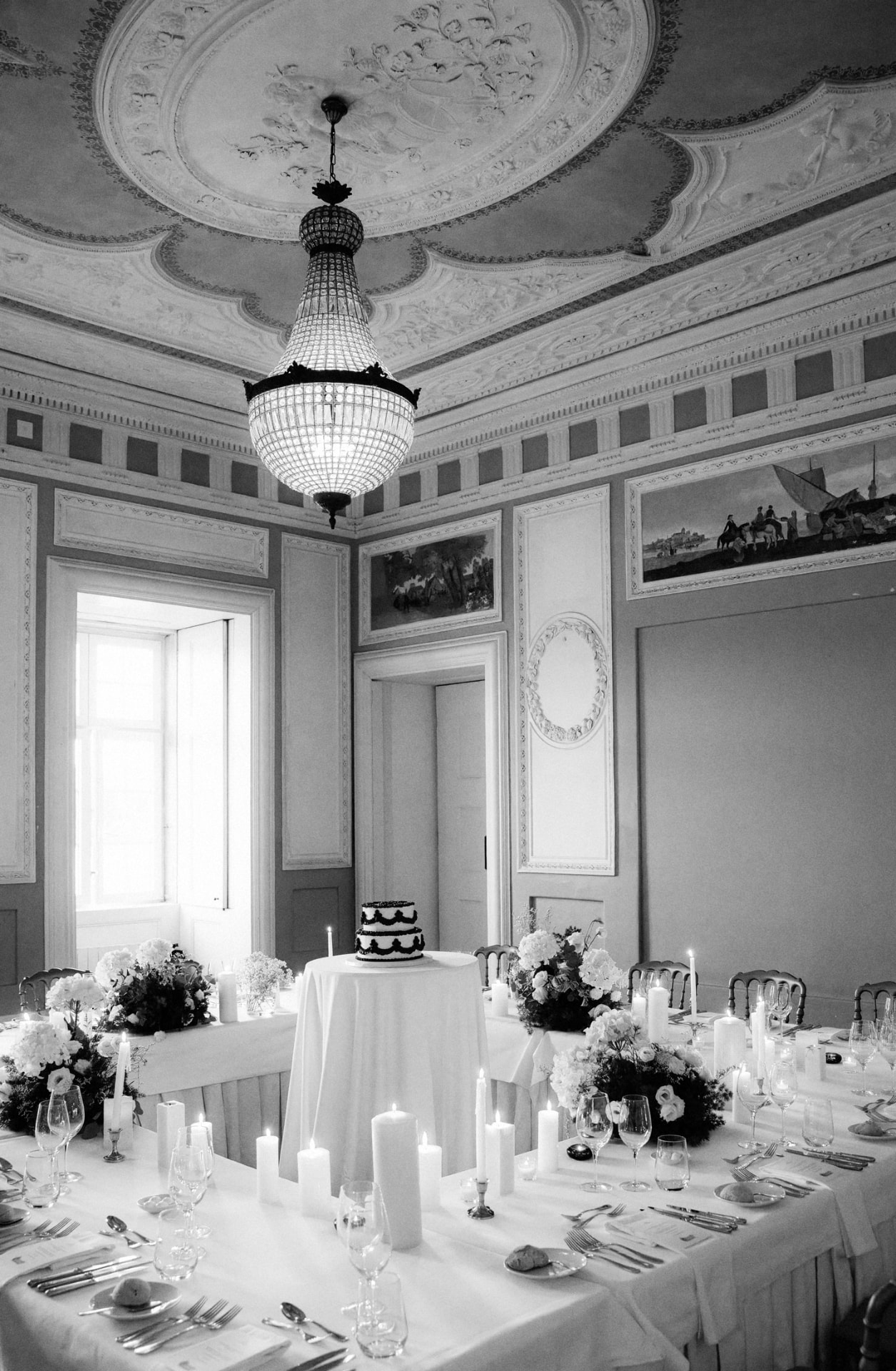 Frescoed palace salon with a crystal chandelier above candlelit banquet tables and a two-tier cake