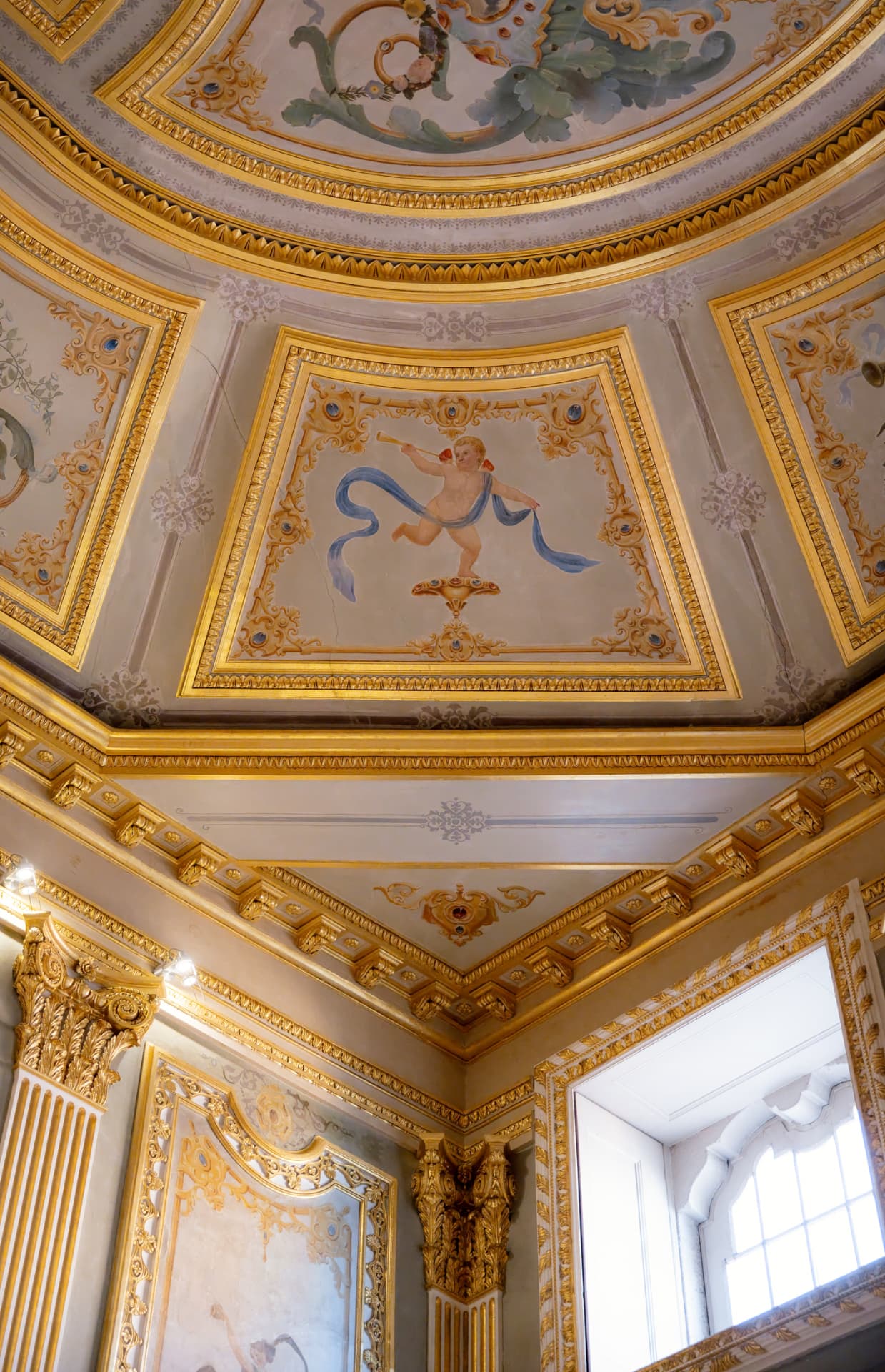 An ornate gilded palace ceiling with painted frescoes and a cherub among gold leaf mouldings