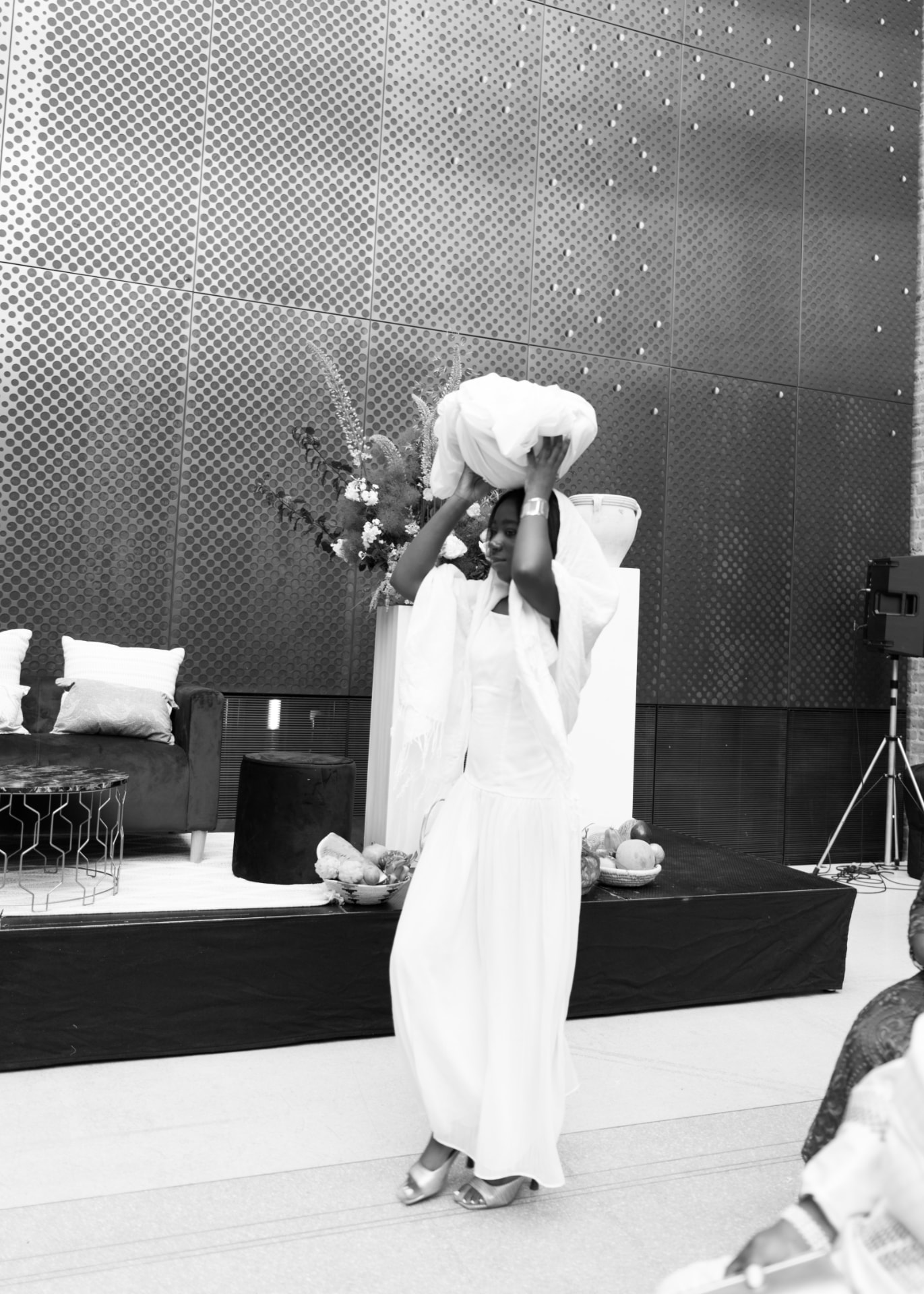 A young woman in a white dress and fringed shawl carries the dowry, wrapped in white cloth, on her head