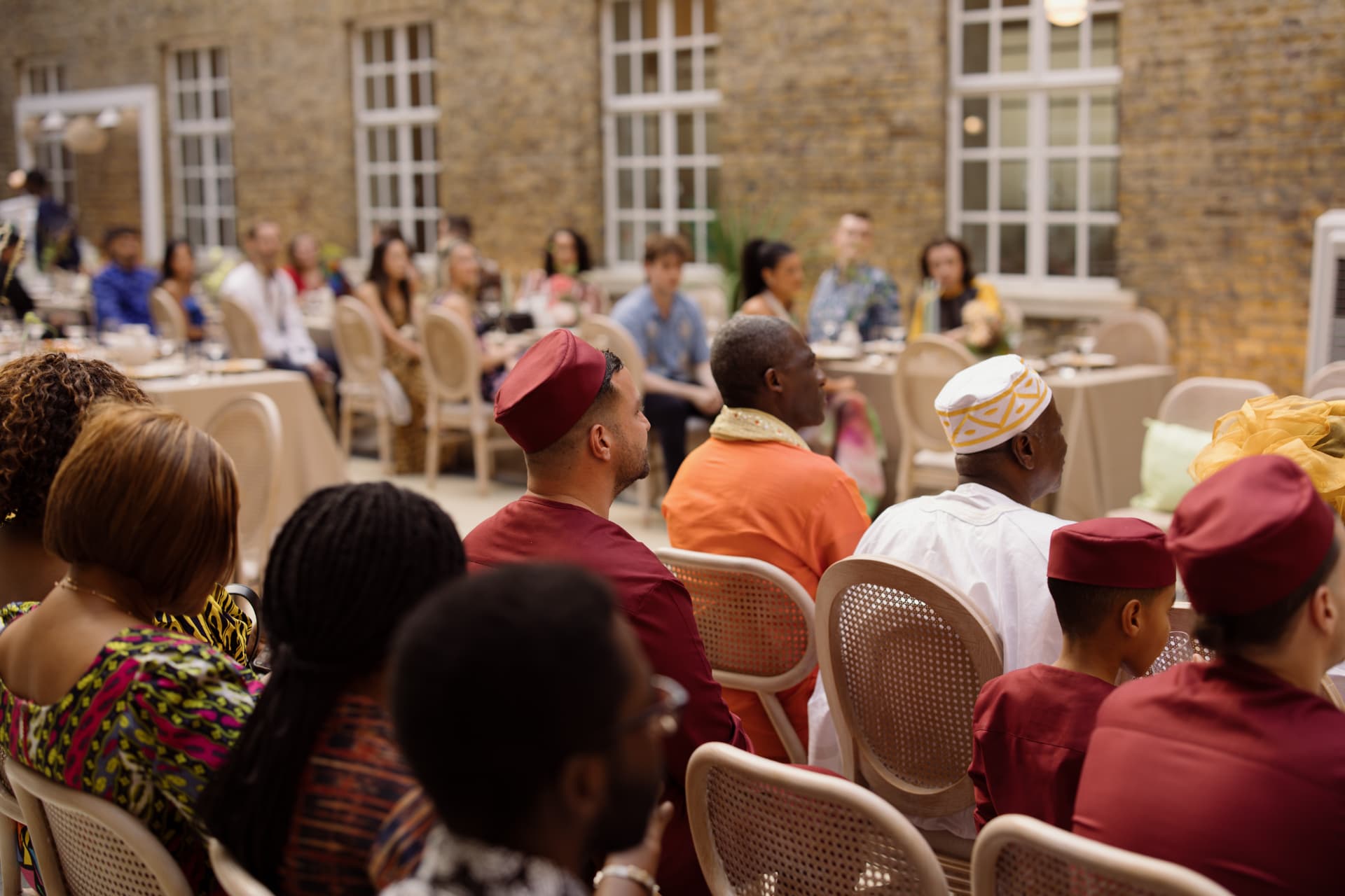 Guests seated at round tables in the courtyard facing the ceremony, elders in the foreground, a man in an orange robe with a gold scarf beside a man in a white tunic and a patterned cap