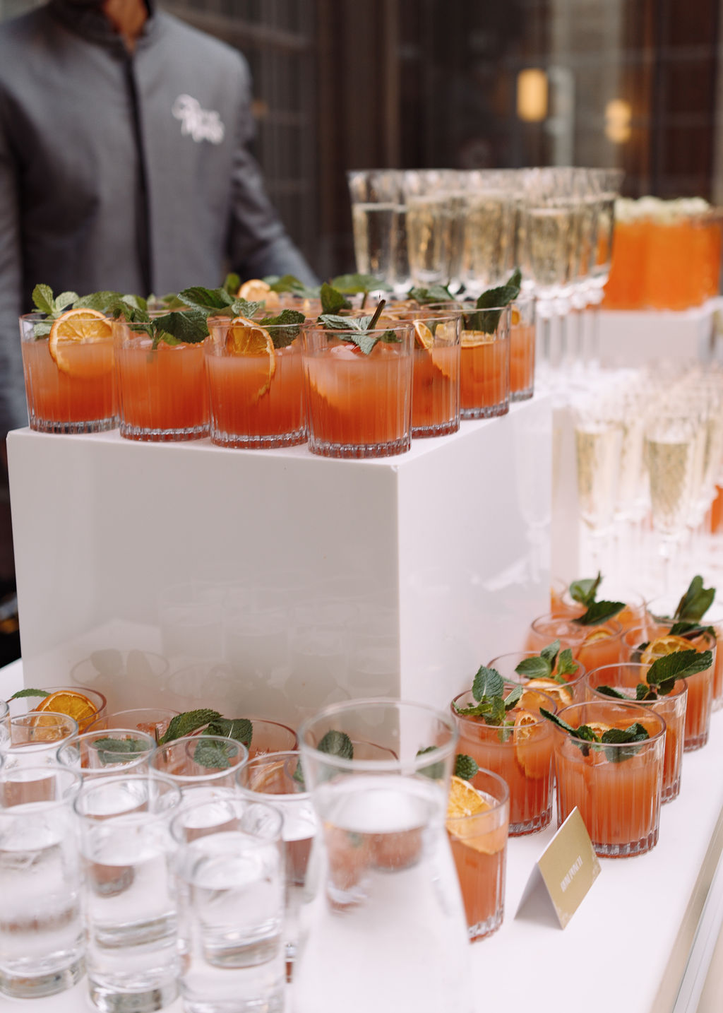 Rows of citrus punch in cut glasses with mint and dried orange, beside champagne flutes on a white display