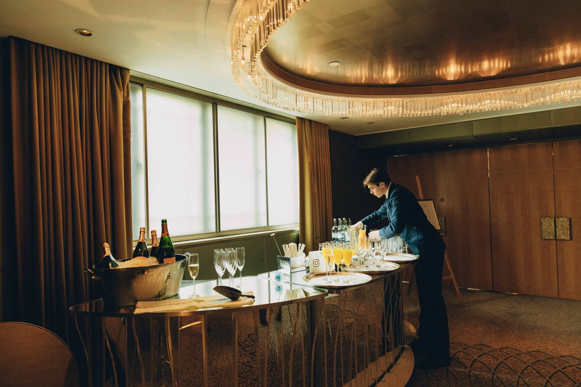 A waiter preparing champagne and juice service on a mirrored bar beside draped windows