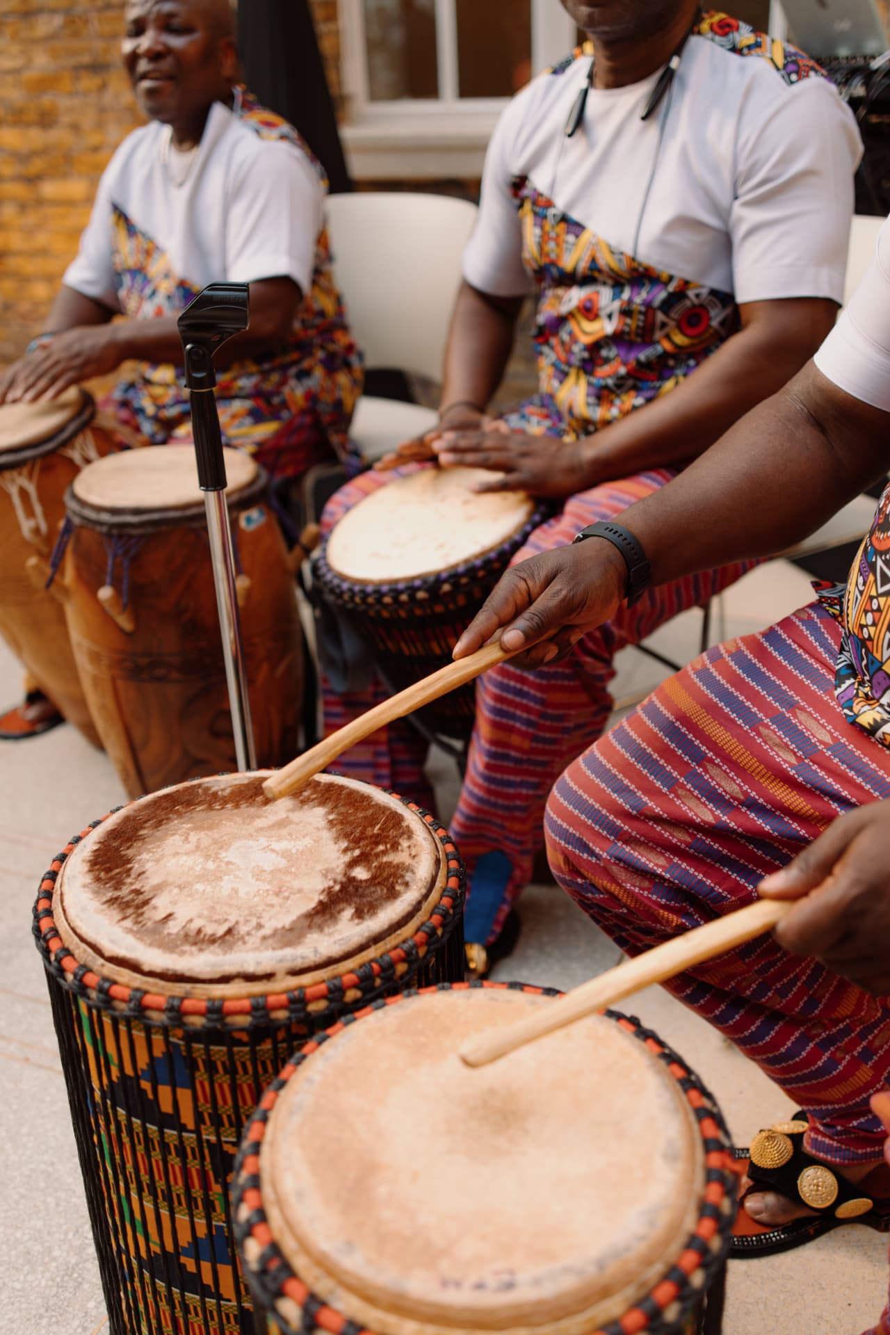 Drummers in patterned cloth playing talking drums in a brick courtyard