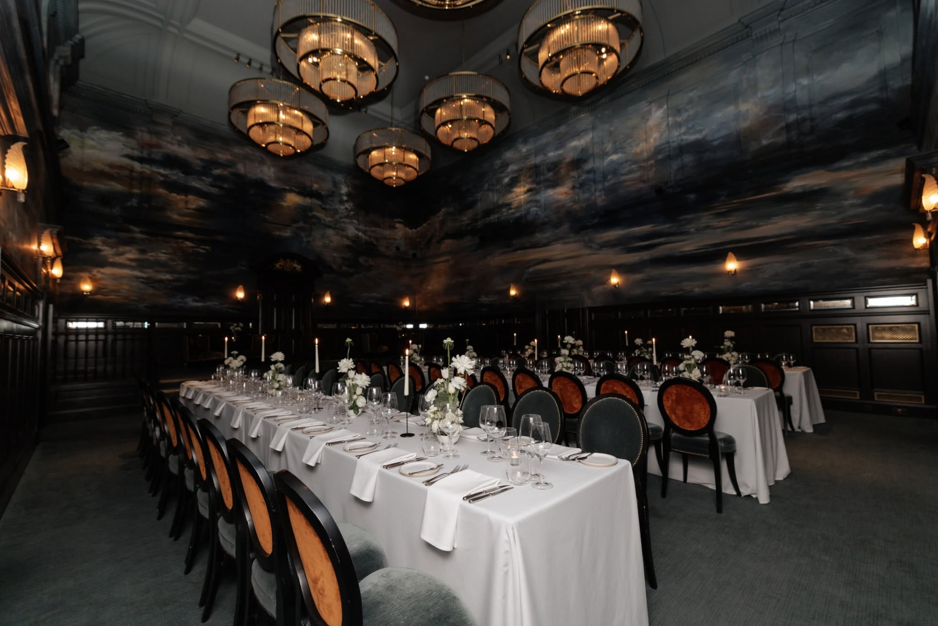 Long banquet table beneath brass chandeliers and painted cloud murals at Nomad Hotel London