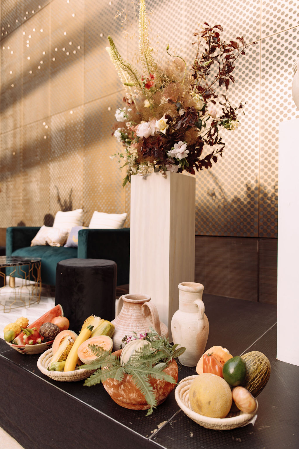 Tropical fruit still-life in woven baskets and terracotta vessels beneath a tall floral arrangement against a perforated brass screen