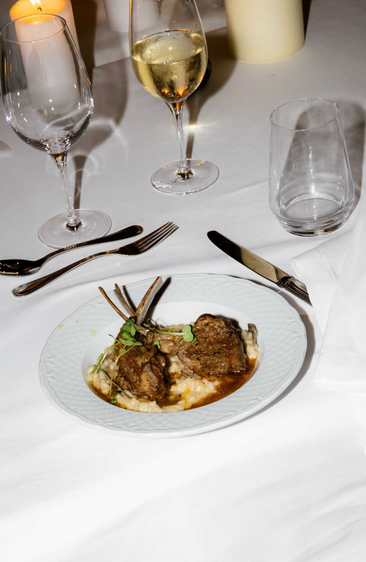 Rack of lamb on risotto plated on porcelain beside crystal glassware and candlelight