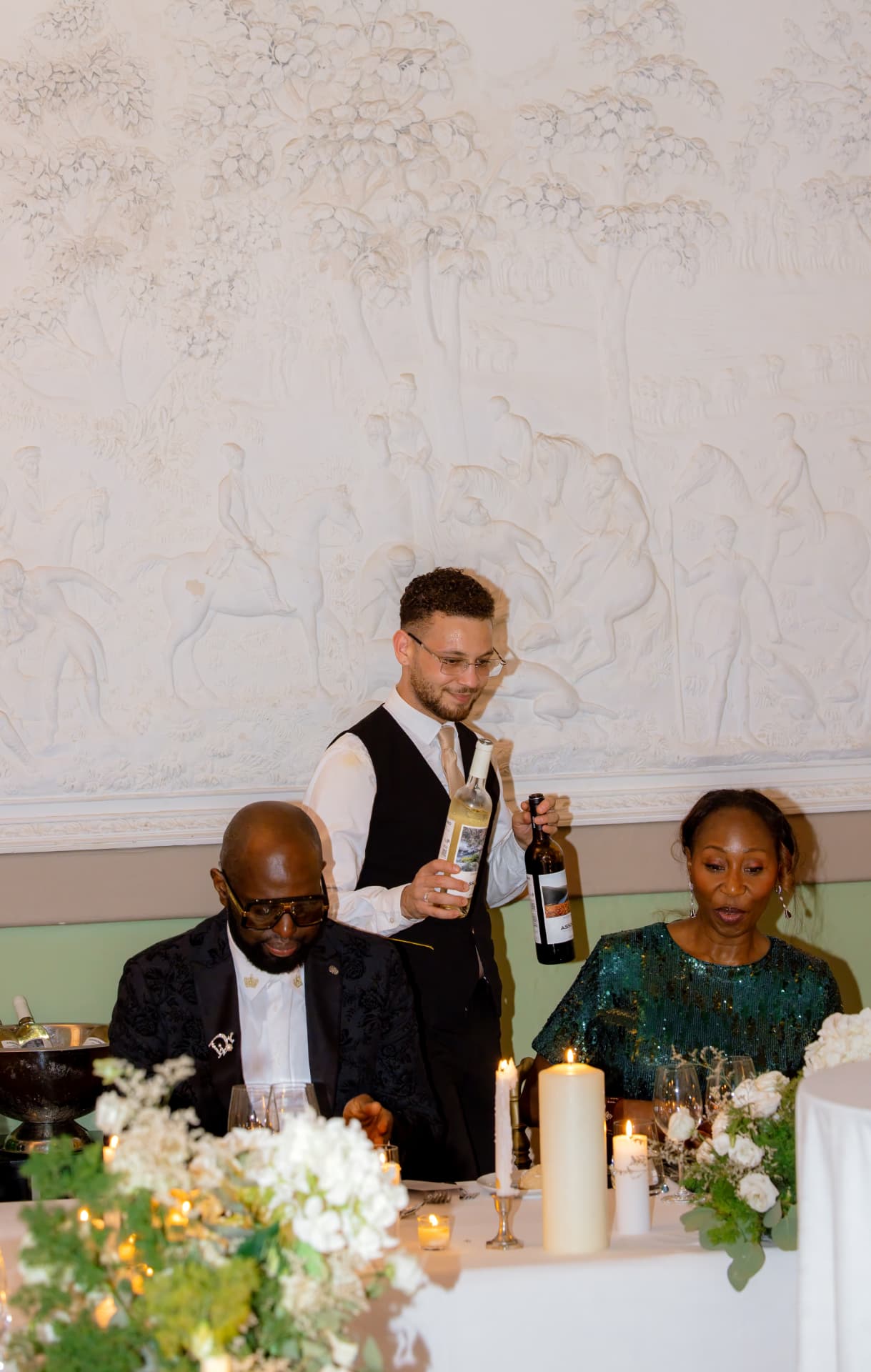 A sommelier presenting two bottles at the top table beneath a carved plaster frieze