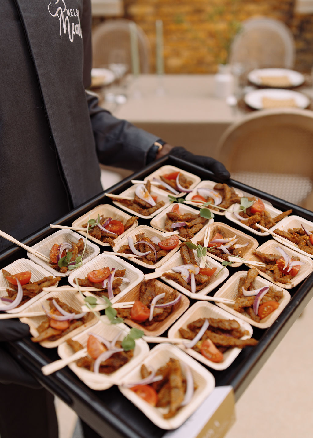 Server carrying a tray of suya canapes with red onion and tomato in palm-leaf dishes
