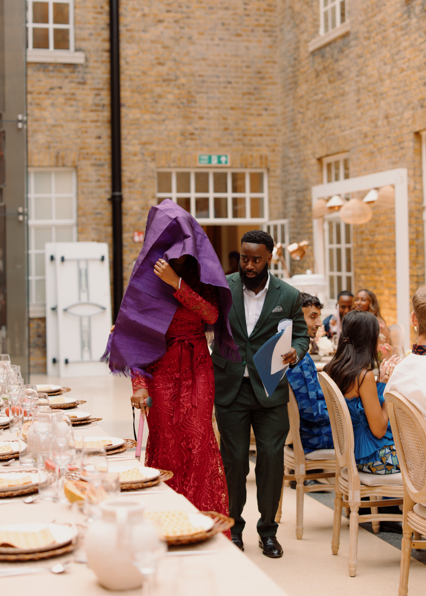 A guest in red lace beneath a purple aso-oke veil walking the length of the banquet table