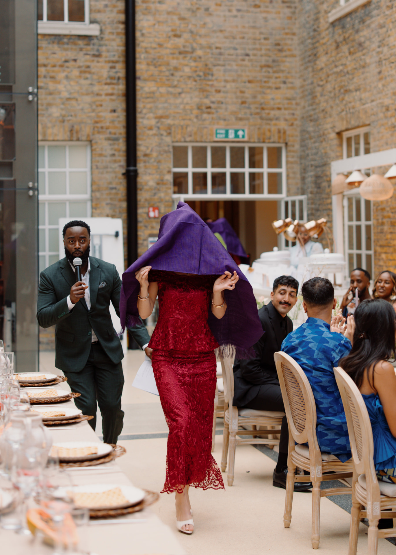 A woman in a burgundy lace dress walks through the courtyard with her head and shoulders completely veiled in a purple cloth, an officiant with a microphone leading the way while guests watch from their tables