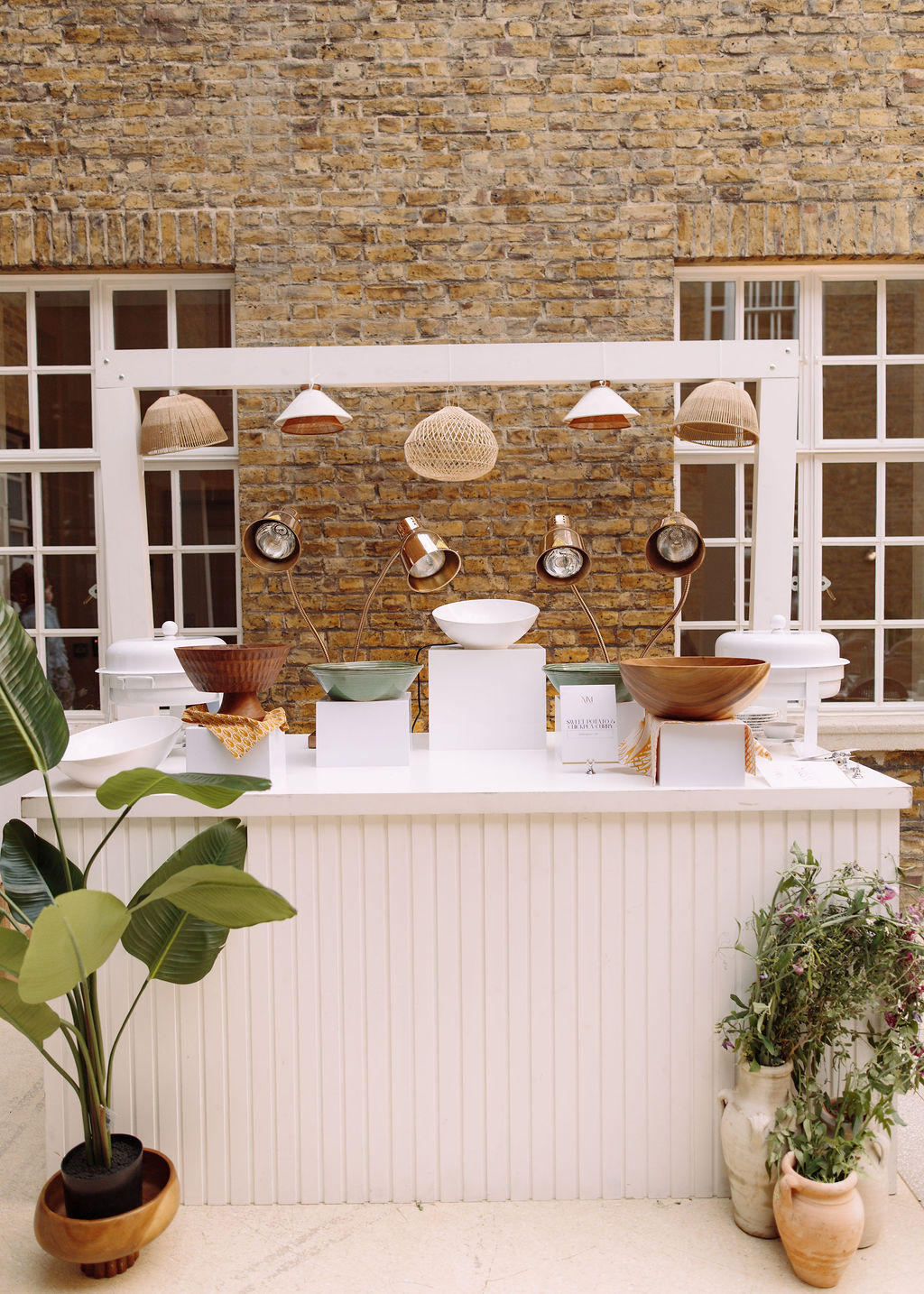White fluted serving station with copper heat lamps, carved wooden bowls and ceramics against a stock brick wall