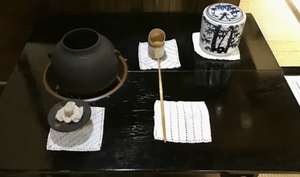 A cast-iron kettle set in a sunken brazier beside a blue and white water jar and a bamboo ladle, laid out for an East Asian tea ceremony