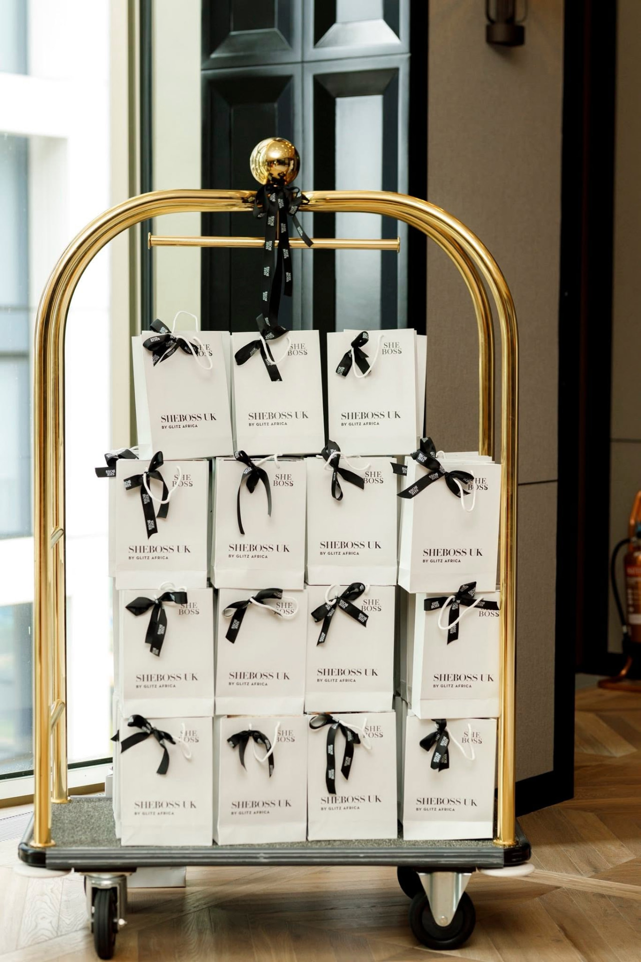 Branded SheBoss UK gift bags stacked on a brass hotel luggage trolley